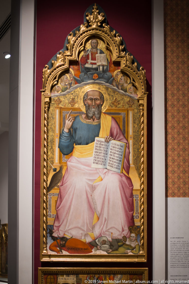 Ornate gilded altarpiece panel with a seated male saint holding an open book and raising one hand.