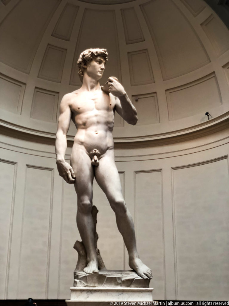 Michelangelo’s marble David standing beneath the Accademia tribune, viewed from below with head turned and sling over his shoulder.