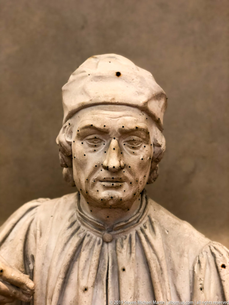 Close view of a worn marble male bust wearing a soft cap and gathered garment, with dark surface marks across the face.