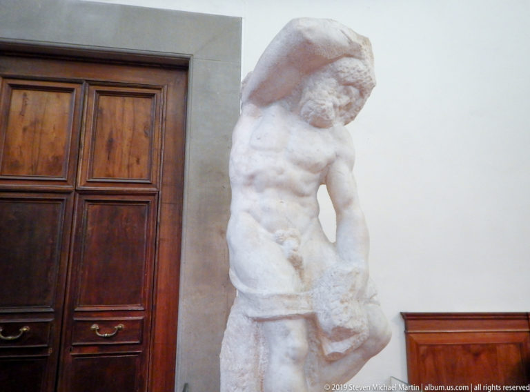 Full-height unfinished Michelangelo captive beside a wooden doorway, with torso emerging below a heavy rough block around the head.