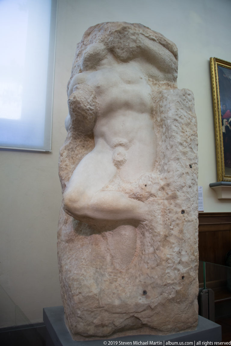 Close side detail of an unfinished Michelangelo captive with chest and head emerging from a thick rough marble block.