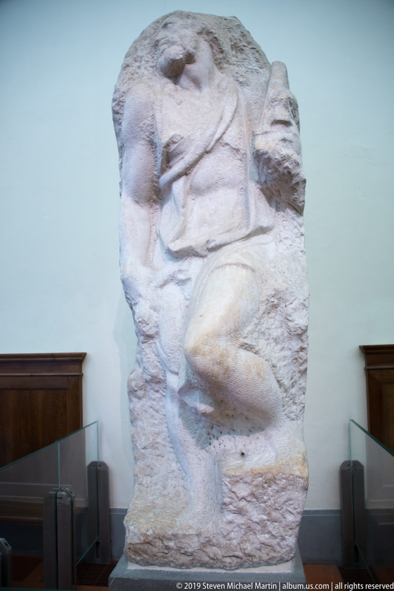 Rough marble block containing a partially emerged Michelangelo captive, with bent limb and partial head visible from the side.