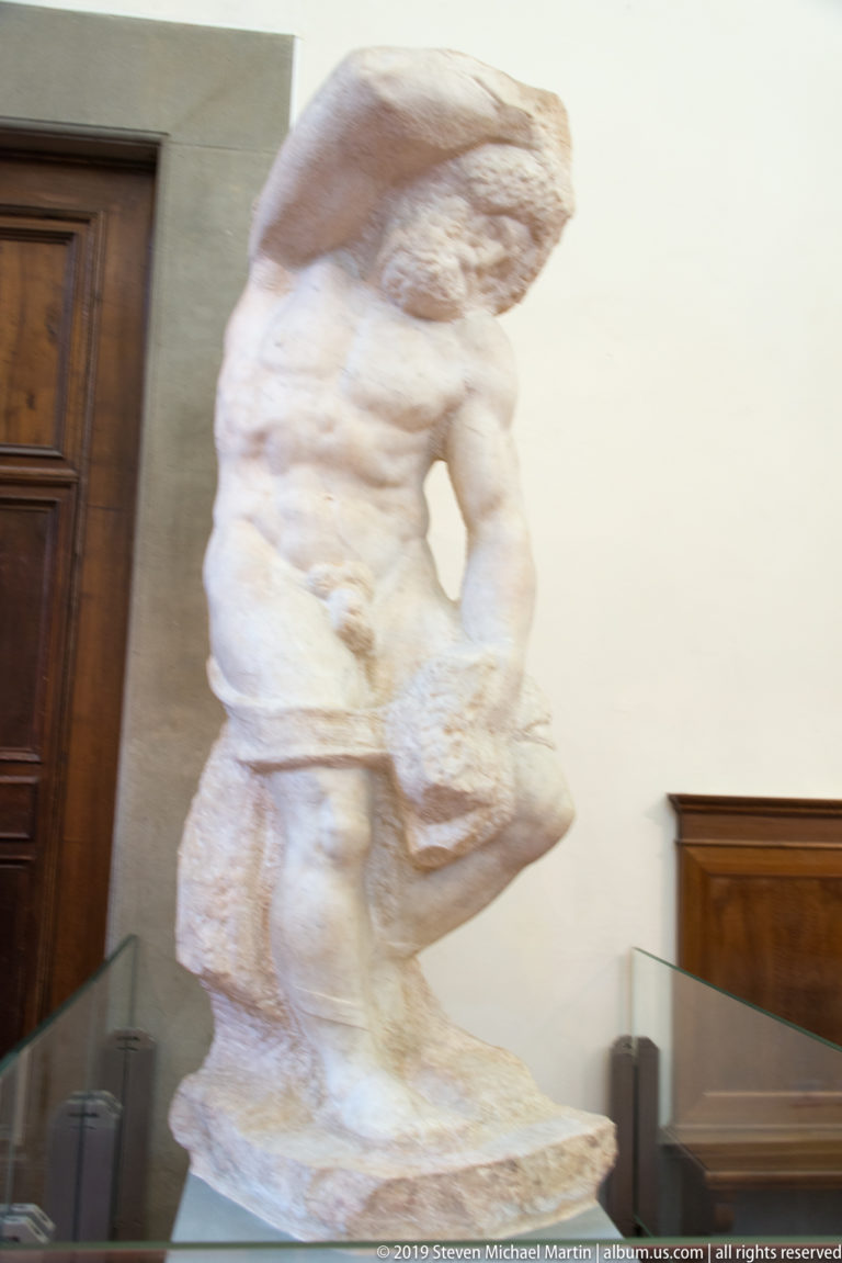 Full view of an unfinished Michelangelo captive emerging from a tall rough marble block beside dark wooden gallery fittings.
