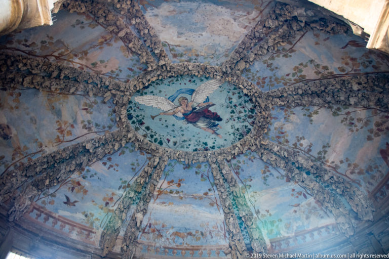 Upward view of an ornate grotto ceiling with central oval painting, radiating encrusted ribs, and pale blue painted compartments.