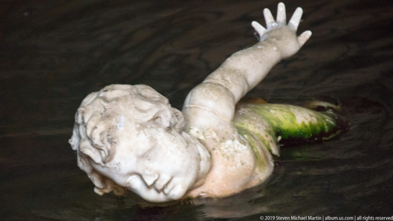 Pale reclining fountain figure partly submerged in dark water, with one arm raised and green staining on the torso.