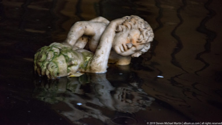 Reclining pale fountain figure resting his head on one hand beside a green-stained vegetal form in dark water.