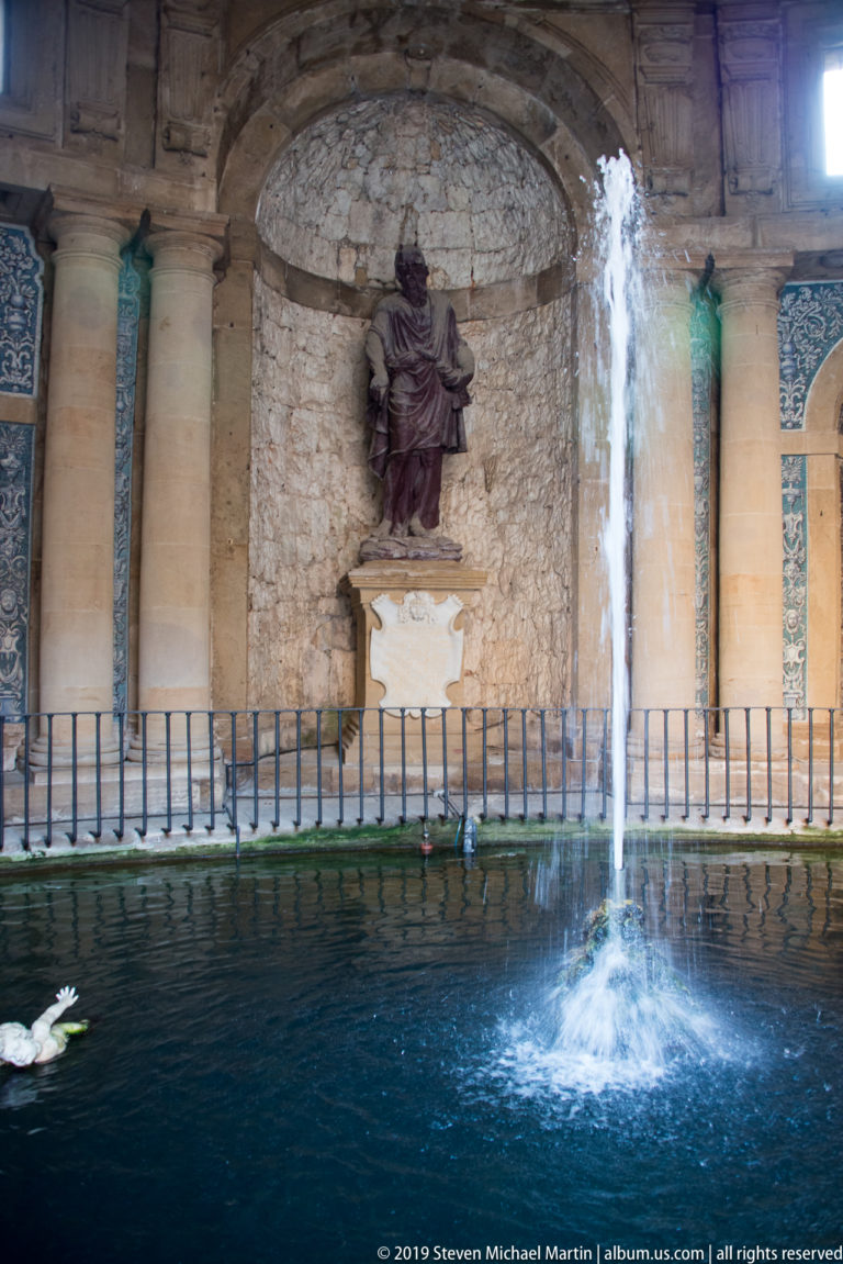 Water jet rising from a dark pool before a statue in a rocky niche, columns, and decorated grotto walls.