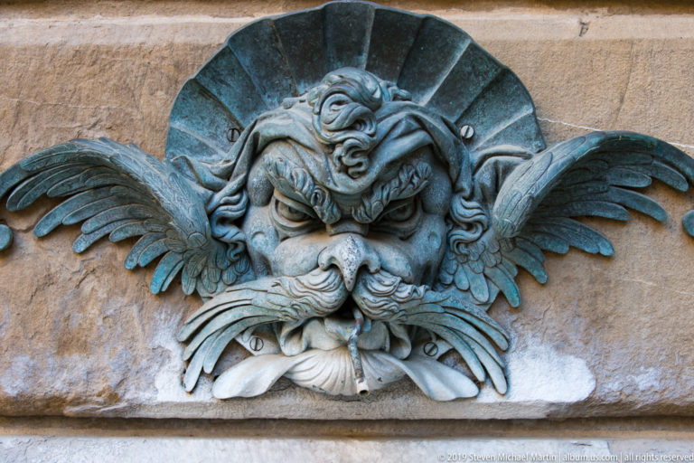 Close view of a weathered winged grotesque face with curling moustache and open mouth mounted on stone.