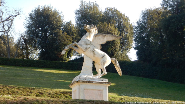 White statue of a rearing winged horse on a pedestal against a grassy slope and trees in the Boboli Gardens.