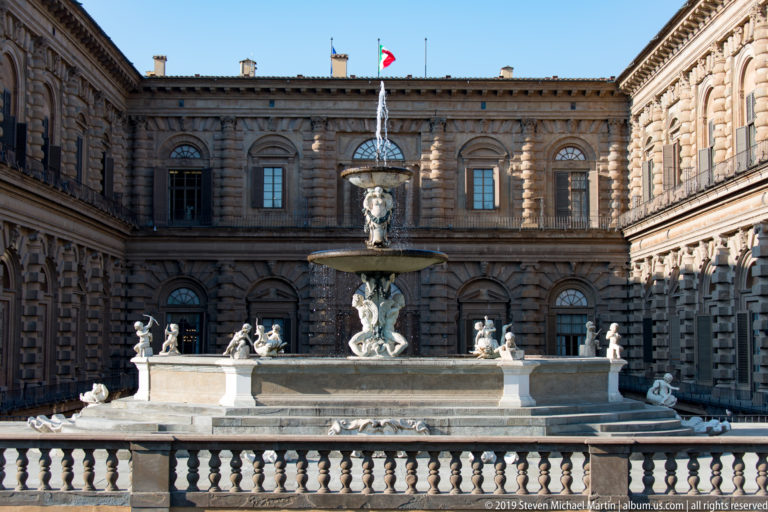 Frontal view of the tiered Artichoke Fountain against Pitti Palace’s rusticated courtyard façade and Italian flag.