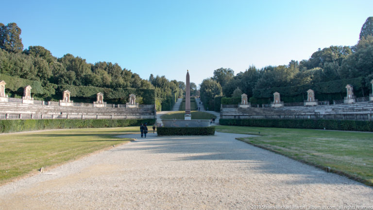Wide gravel path and lawn leading to an Egyptian obelisk, with curved statue-lined terraces and trees on both sides.
