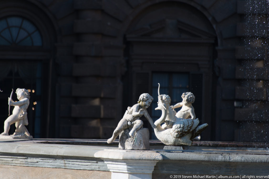 Sunlit small sculpted figures along a fountain basin, silhouetted against a dark arched doorway in Pitti Palace.