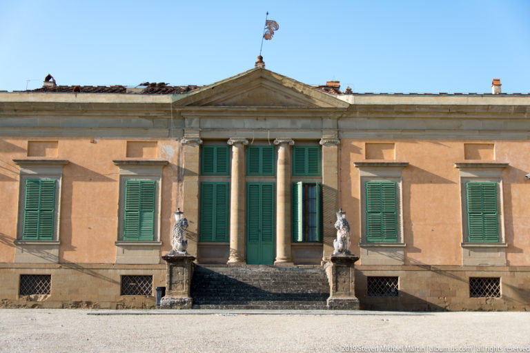 Symmetrical ocher garden pavilion with green shutters, central columns and pediment, broad steps, and statues flanking the entrance.