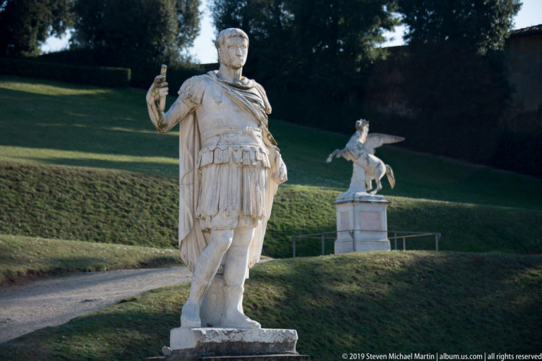 Marble statue of an armored Roman-style male figure beside a garden path, with a winged horse sculpture in the background.