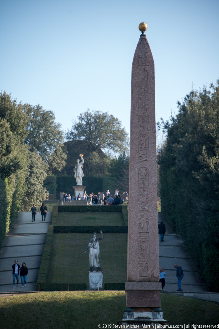 Tall hieroglyph-covered Egyptian obelisk centered among terraces, hedges, statues, and visitors in the Boboli Gardens amphitheater.