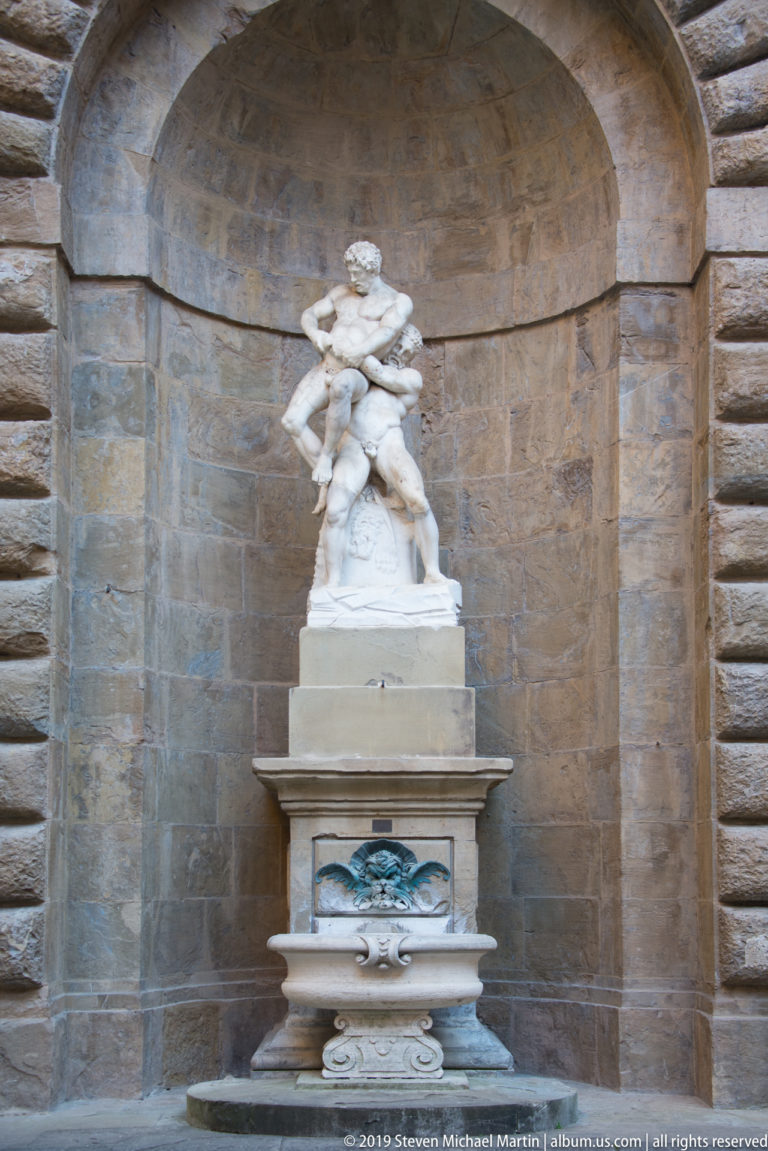 Small white marble figure group above a projecting basin in a large arched niche of rusticated stone.