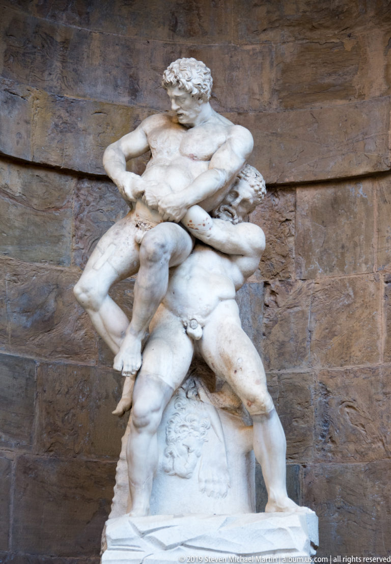 White marble sculpture of two nude male figures locked in a physical struggle against a rusticated stone wall.
