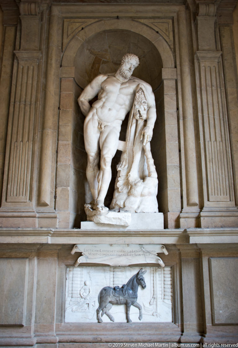 Large white marble sculptural group of a muscular nude supporting another figure in a deep rusticated stone niche.