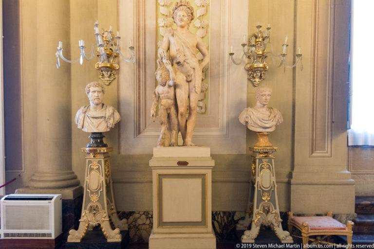 Long decorated palace gallery with sculpture displays, chandeliers, and architectural ornament.