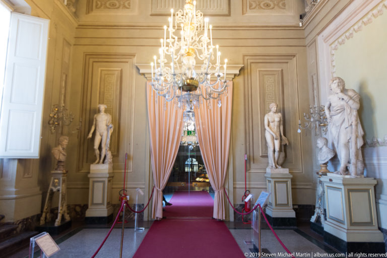 Long decorated palace gallery with sculpture displays, chandeliers, and architectural ornament.