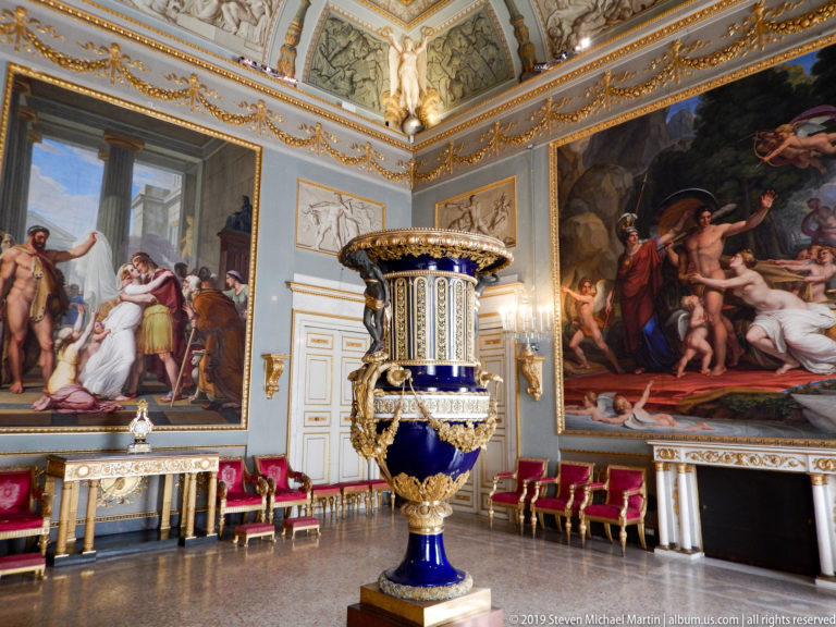 Long decorated palace gallery with sculpture displays, chandeliers, and architectural ornament.