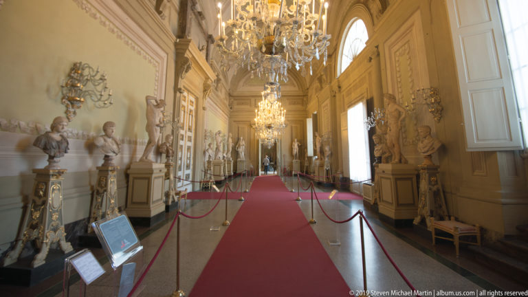 Long decorated palace gallery with sculpture displays, chandeliers, and architectural ornament.