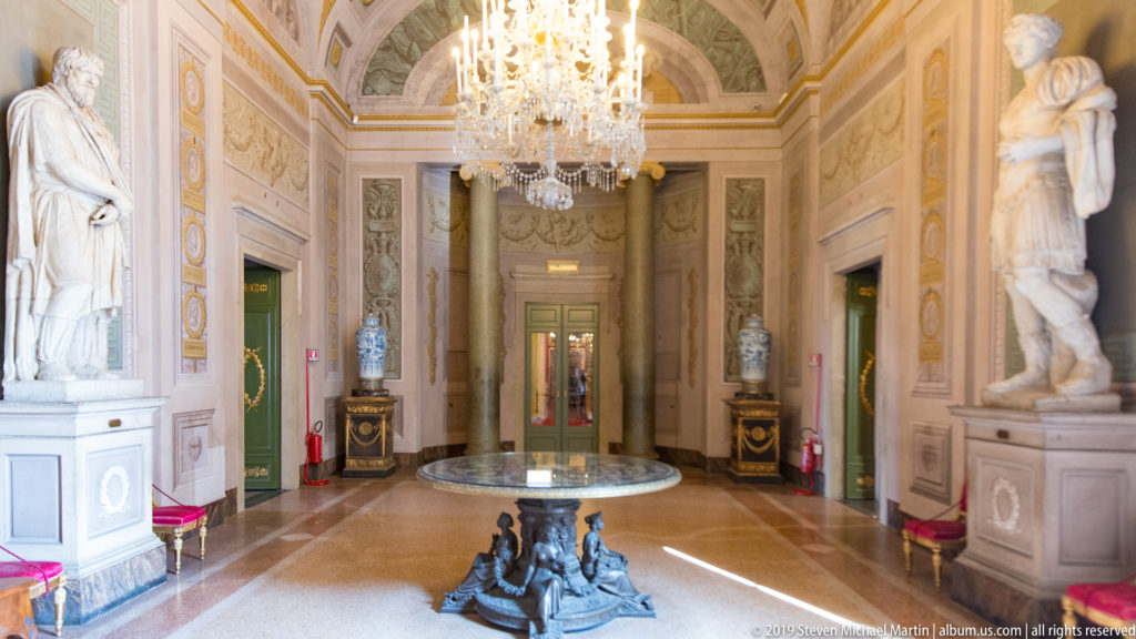 Long decorated palace gallery with sculpture displays, chandeliers, and architectural ornament.