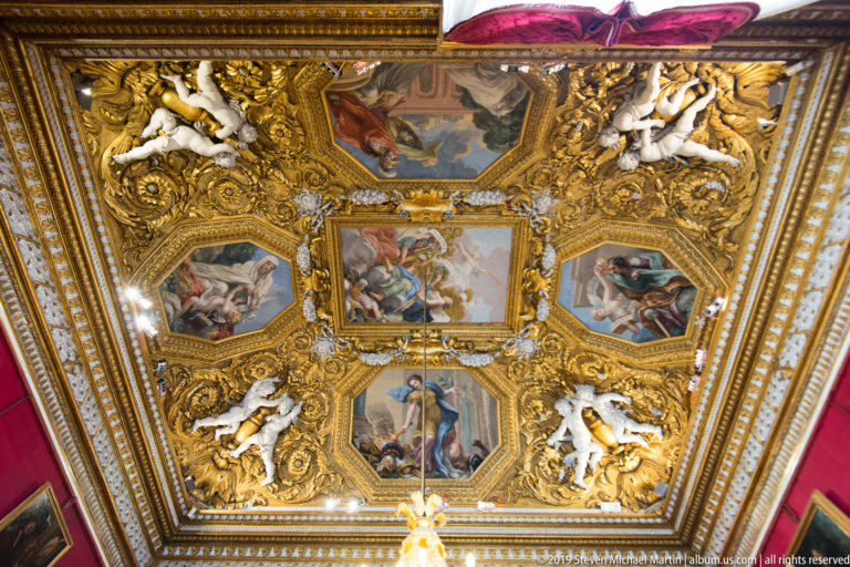 Large framed or ceiling painting with multiple figures in a richly decorated Pitti Palace interior.