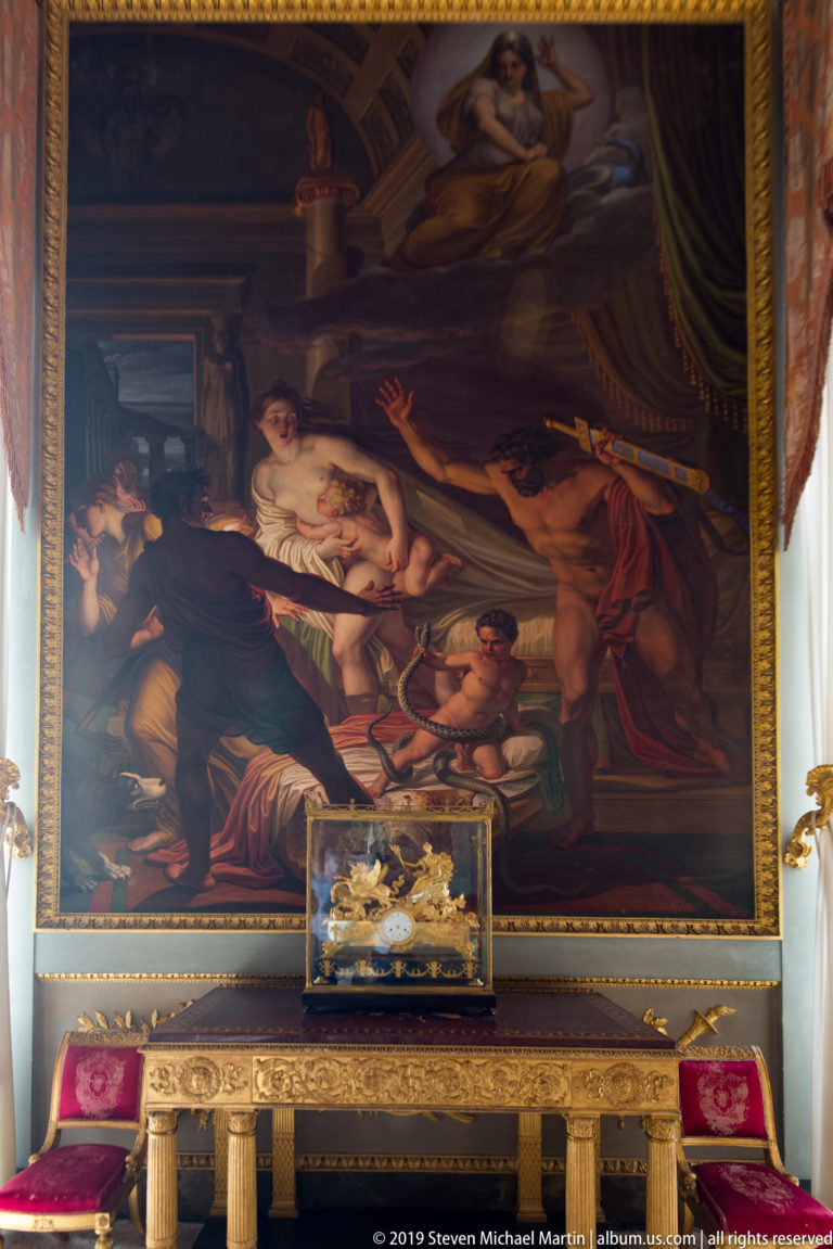 Large dark multi-figure painting above an ornate console and decorative casket, flanked by red upholstered chairs.
