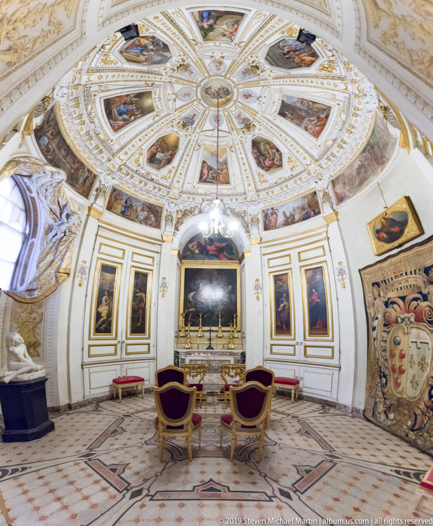 Circular palace room with radial frescoed dome, white-and-gold walls, framed paintings, patterned marble floor, and paired chairs.