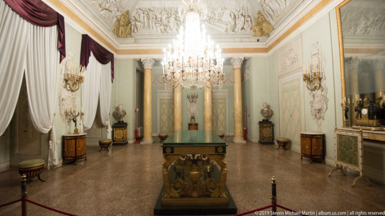 Pale green neoclassical room with paired columns, crystal chandelier, busts, wall lights, drapery, and polished furniture.
