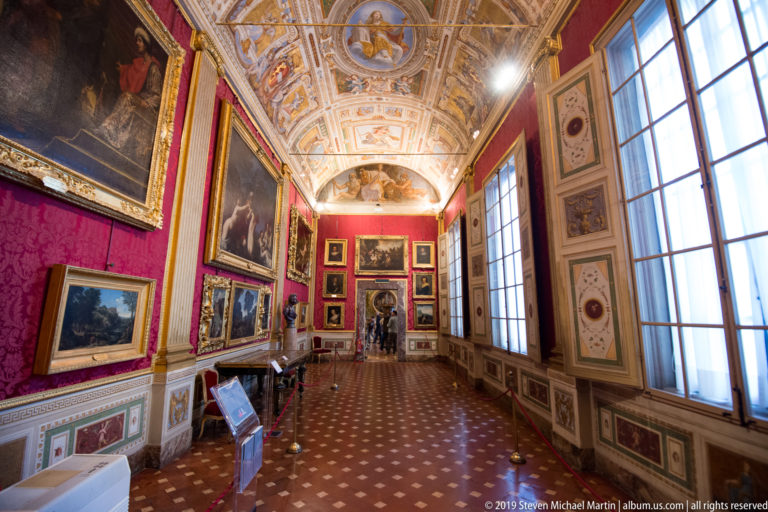 Long red-walled gallery lined with paintings, tall windows, patterned floor, decorated ceiling, and a distant doorway.