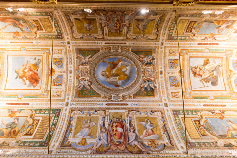 Elaborately compartmented ceiling filled with grotesque ornament, painted figures, heraldic shields, and a central round scene.
