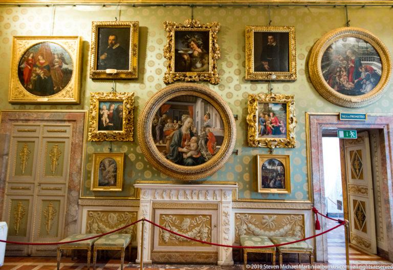 Historic gallery room with densely hung framed paintings, patterned walls, carved mantel, gilded furniture, and a roped visitor barrier.
