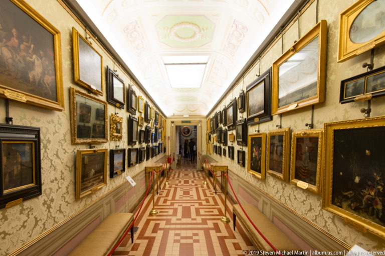 Narrow bright corridor densely hung with framed paintings on both walls, with red ropes and a geometric patterned floor.