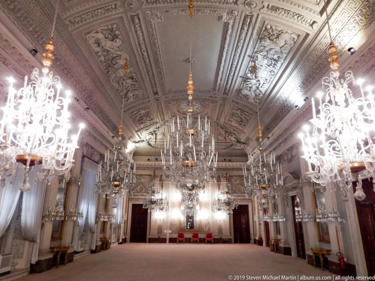 Oblique view across the White Hall at Pitti Palace, with crystal chandeliers, mirrors, pale walls, and elaborate Neoclassical ceiling stuccowork.