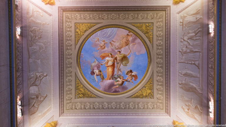 Oval colorful mythological ceiling fresco surrounded by gray relief-like panels and small gilded corner ornaments.
