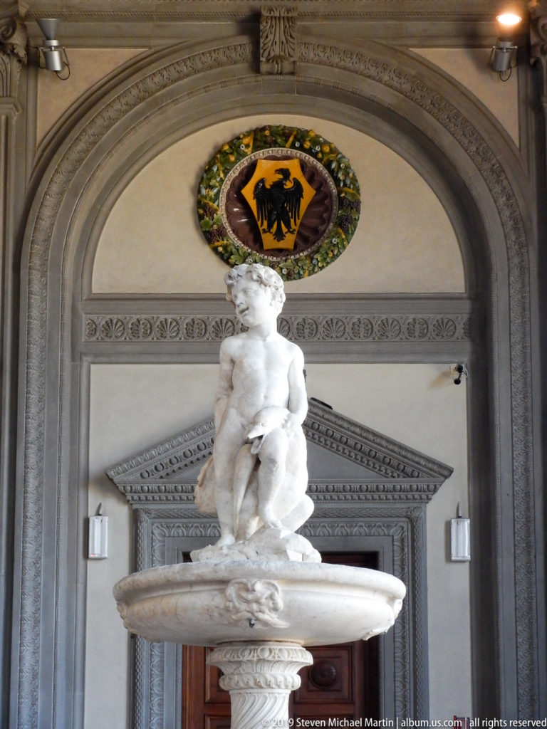 White marble putto fountain centered before a gray arched doorway, with a colorful heraldic roundel above.
