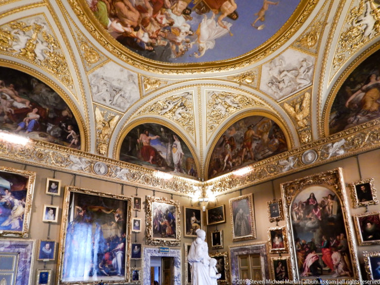 Frescoed and gilded vault above a gallery densely hung with paintings and a white sculpture at floor level.