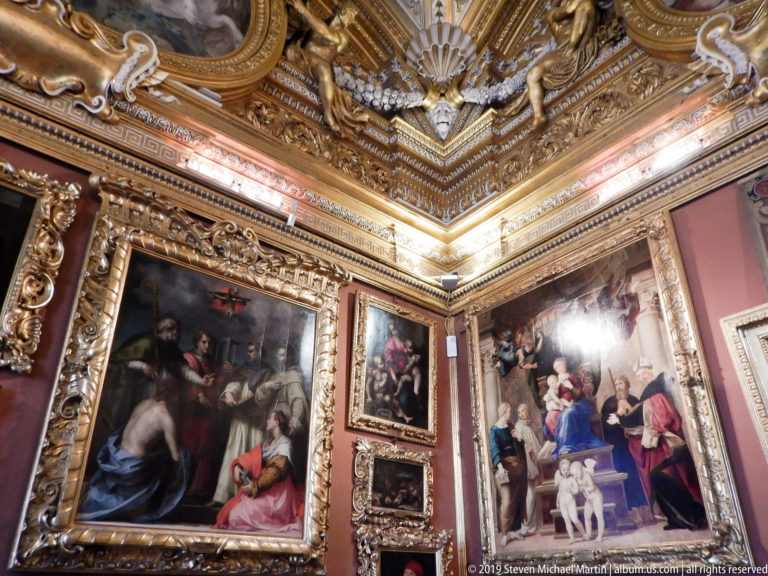 Corner of red-walled gallery with large framed paintings beneath a heavily gilded cornice with shell and figure motifs.