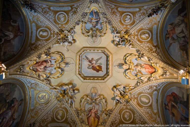 Symmetrical ornate ceiling with central painted panel, gilded stucco cartouches, relief figures, and curved fresco compartments.