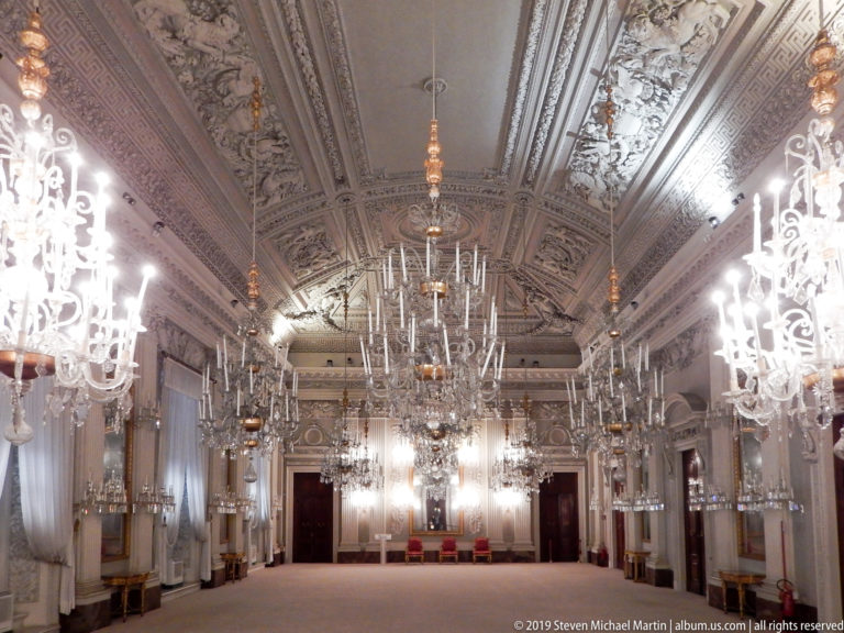 Long axial view of Pitti Palace’s White Hall with elaborate pale stuccowork, mirrors, tall windows, and rows of crystal chandeliers.