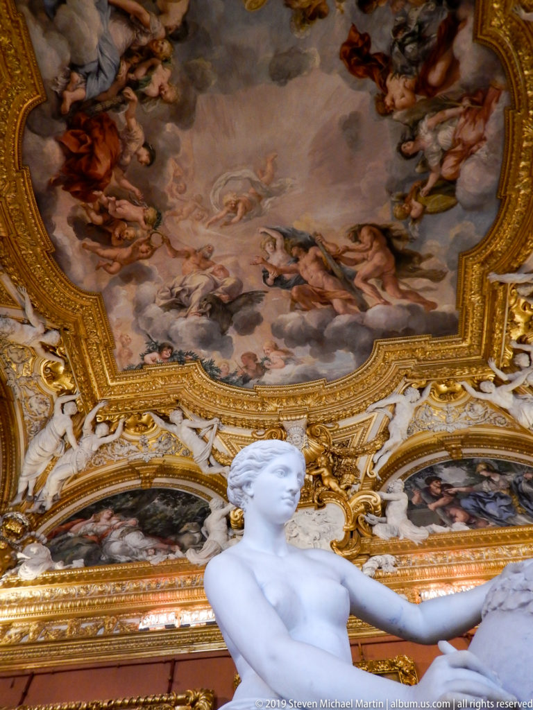 Upper half of a white female statue seen from below against an ornate frescoed and gilded ceiling.