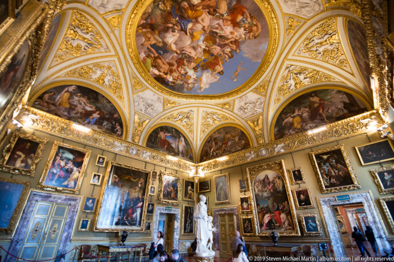 Wide ornate gallery with paintings, white sculpture, visitors, patterned floor, and frescoed vaulted ceiling.