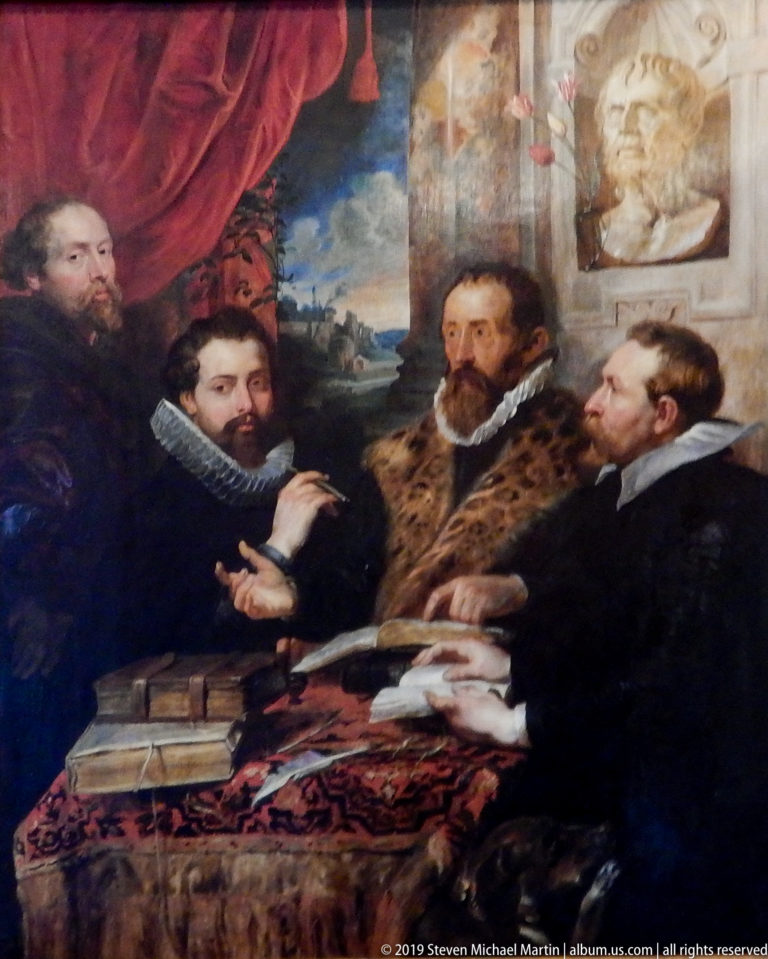 Four bearded and dark-haired men gathered around books and papers beside a classical bust and red drapery.