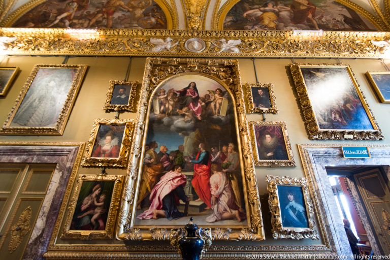 Large religious oil painting in a gilded frame surrounded by smaller paintings beneath an ornate decorated ceiling.