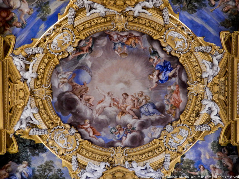 Oval mythological ceiling fresco surrounded by gilded scrollwork, white stucco figures, and blue-painted corner scenes.