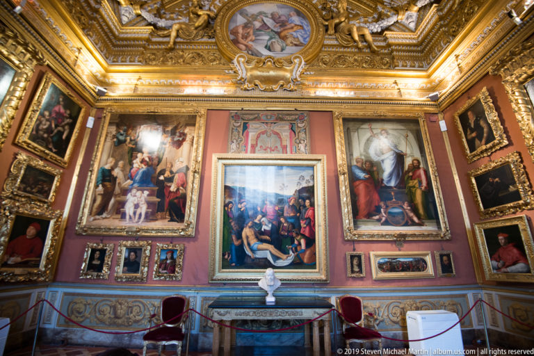 Ornate gallery room with rose walls densely hung with paintings beneath a gilded ceiling and painted medallion.