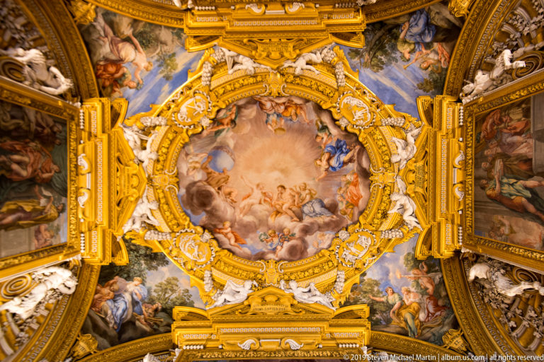 Round mythological fresco of airborne figures and clouds enclosed by elaborate gilded frame with white stucco figures.