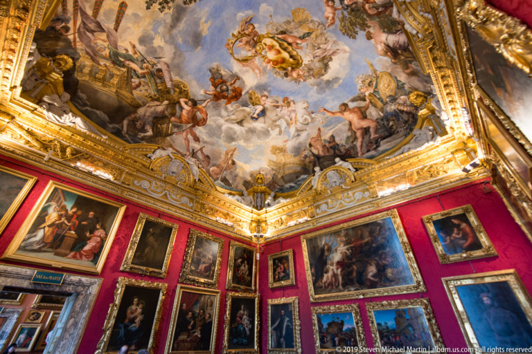 Gilded ceiling and painted figures rise above red walls densely covered in framed paintings, seen toward a corner.